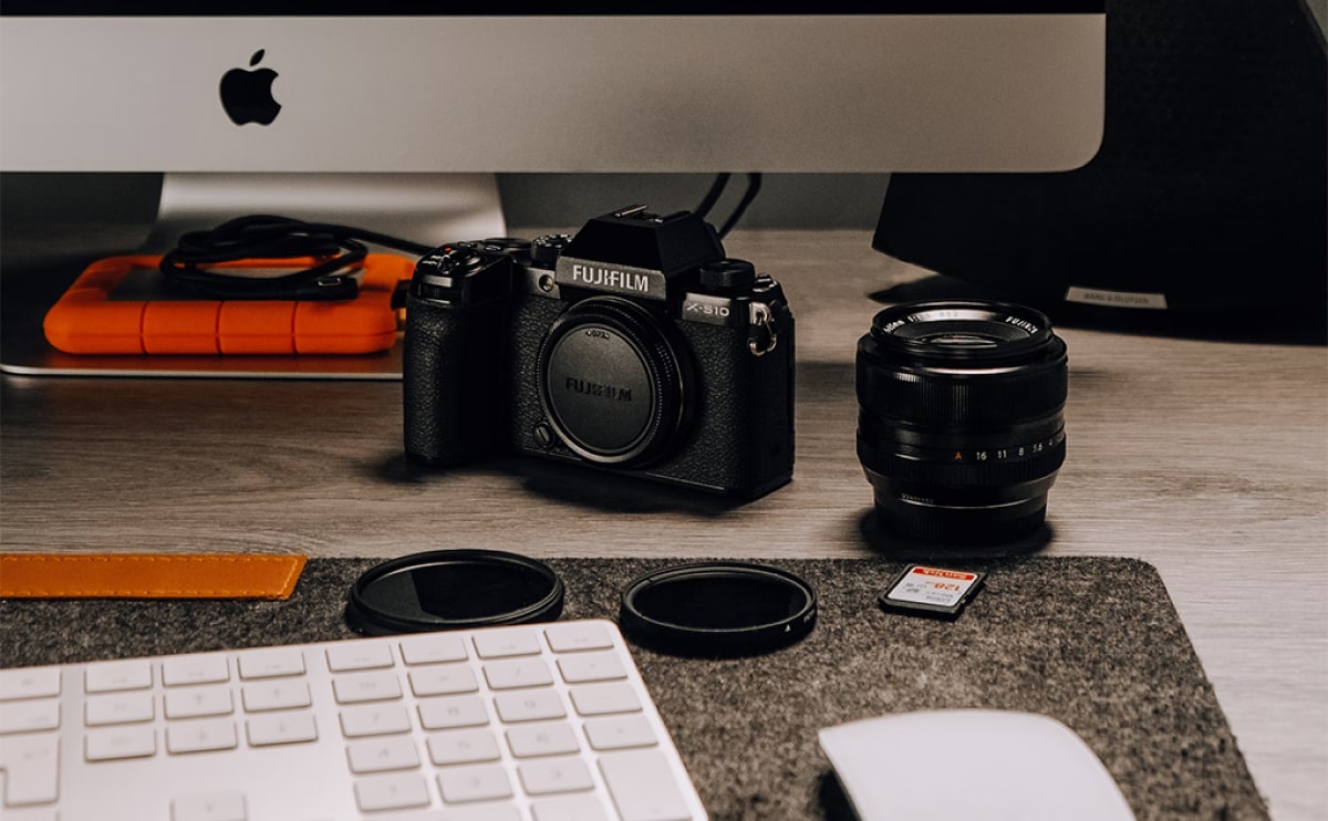 Un appareil photo Fujifilm avec un objectif et un capuchon détachés se trouve sur un bureau à côté d'un imac, d'un clavier et d'un disque dur externe orange. une carte mémoire est également visible sur la table.